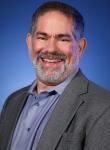 Bearded Joel Sacks, a man, smiling wearing a gray suit and blue shirt